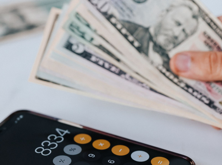 Hand holding US banknotes beside a smartphone calculator.