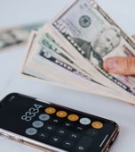 Hand holding US banknotes beside a smartphone calculator.