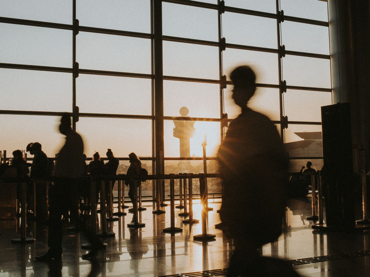Travelers silhouetted against terminal windows in warm sunlight.